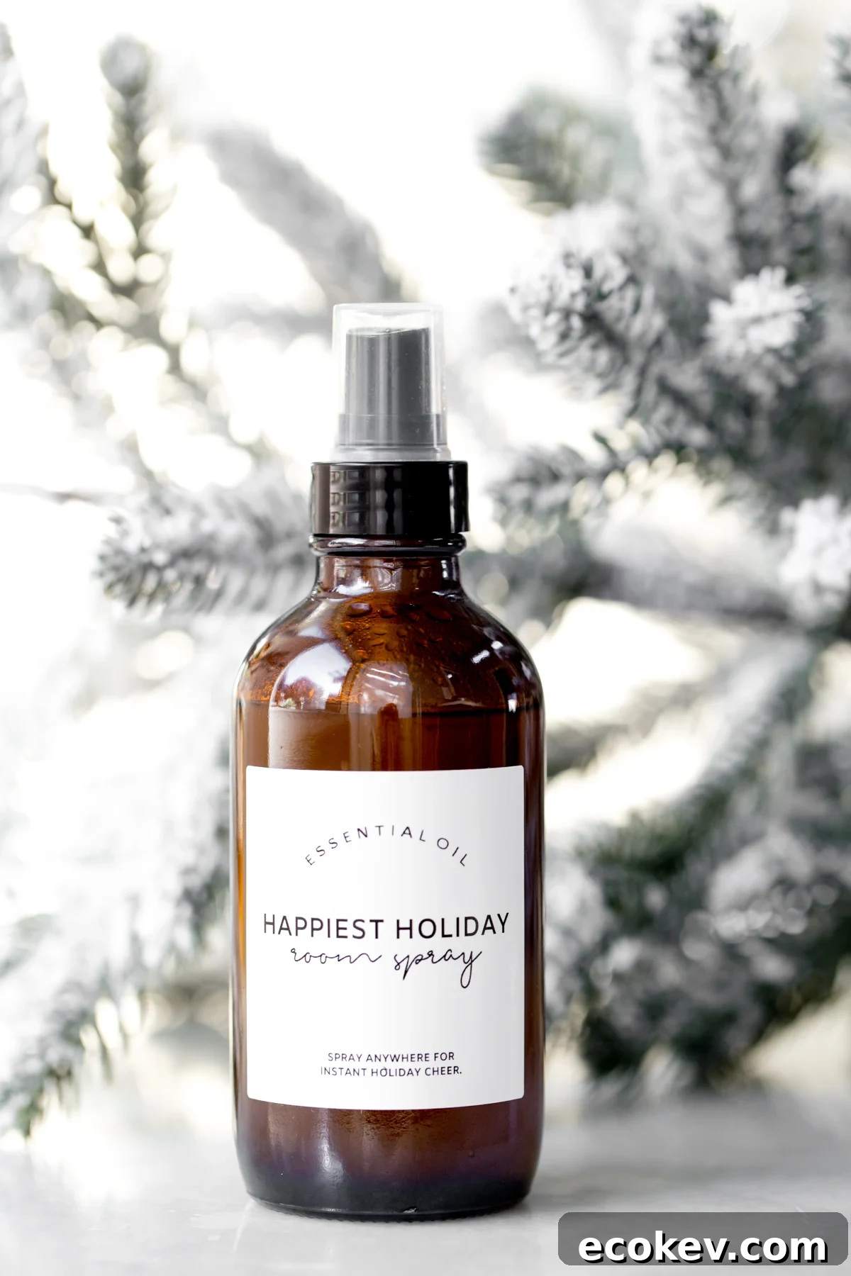 Homemade essential oil Christmas room spray on marble with snow covered tree branches in the background