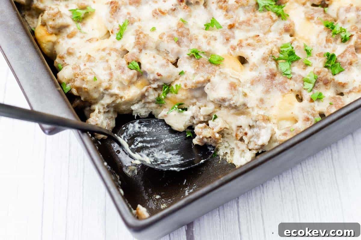 A baking dish of sausage and biscuit casserole topped with creamy gravy and chopped parsley, with a serving spoon partially in the dish.