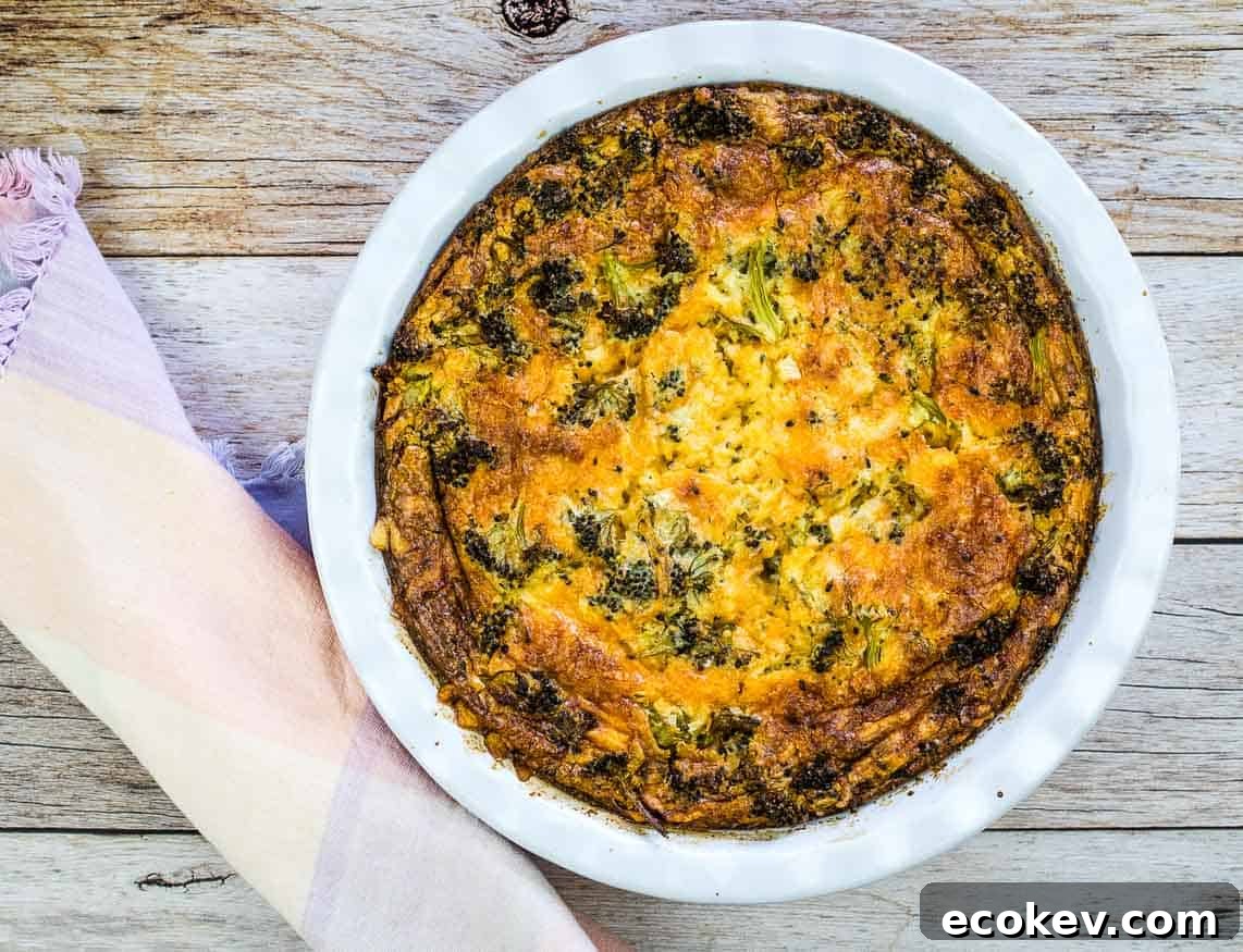 A slice of Broccoli 3-Cheese Impossible Quiche on a white plate with a side of mixed fruit. A full quiche and glass of orange juice are in the background.
