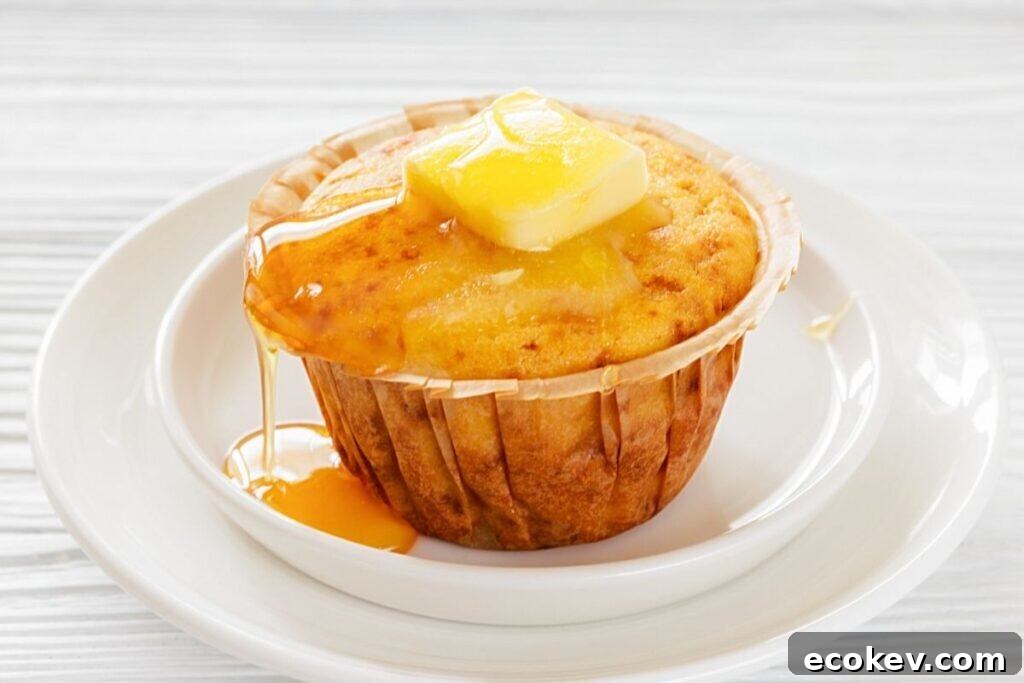 A Honey Cornbread muffin on a white plate, topped with a pat of butter and drizzled with syrup.