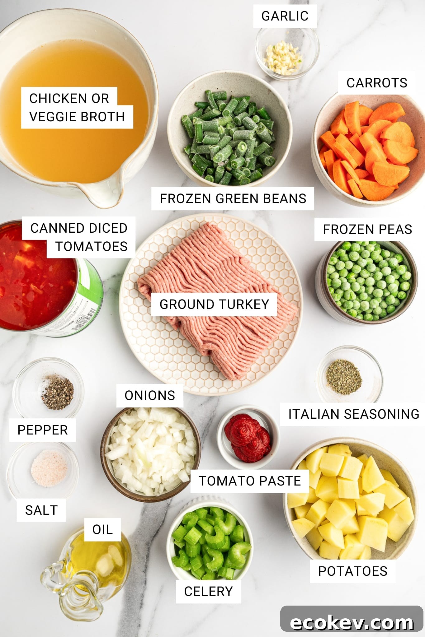 Overhead ingredients photo on a white marble surface with white text labels on each item: broth, garlic, carrots, green beans, peas, ground turkey, onions, tomato paste, Italian seasoning, potatoes, celery, pepper, salt, oil, and canned diced tomatoes.