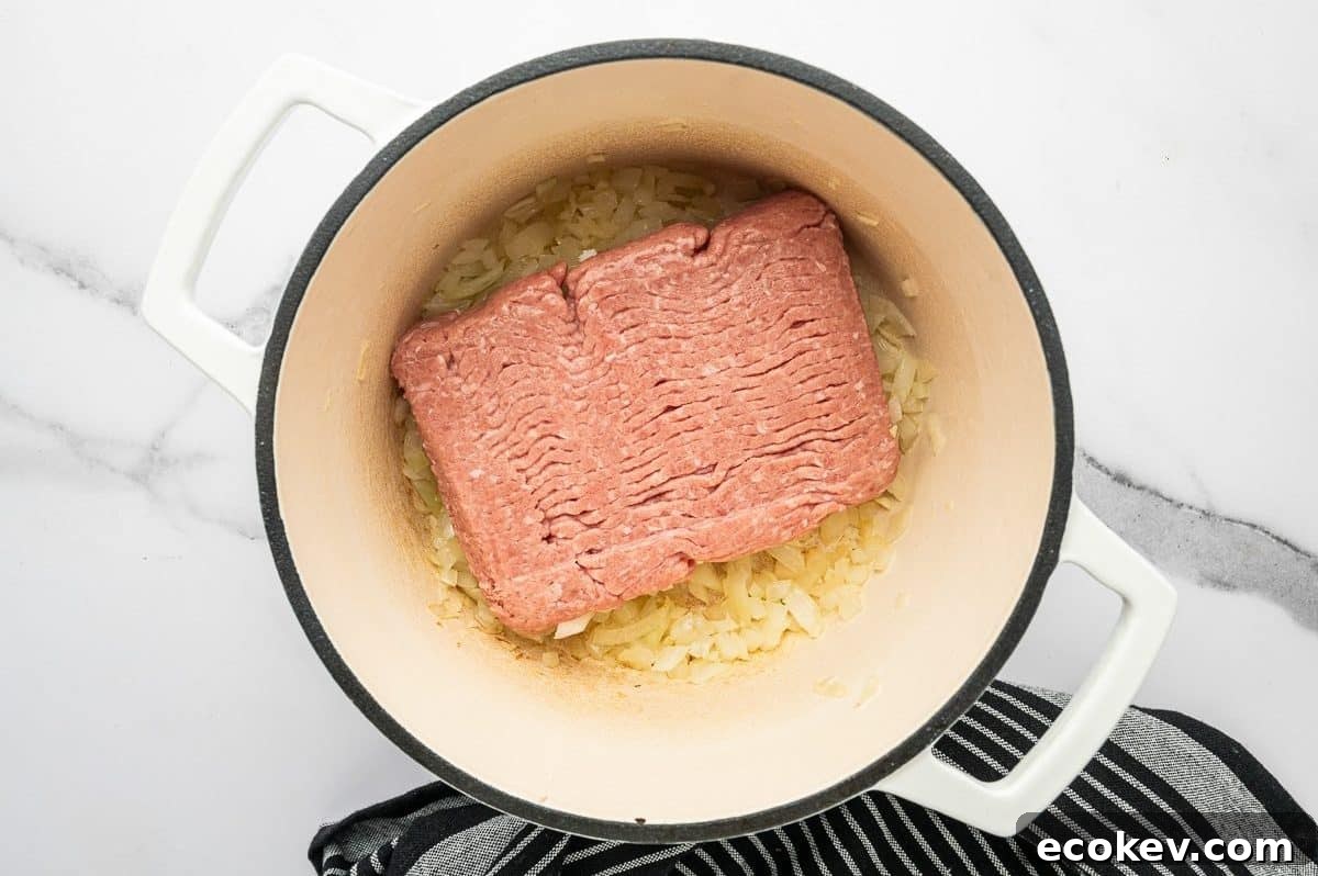 Overhead photo of a block of raw ground turkey placed on top of softened chopped onions and garlic in a white enameled Dutch oven.