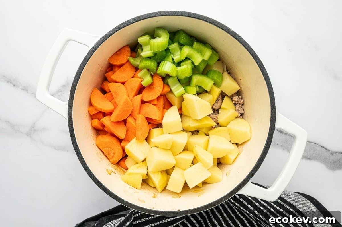 Overhead photo of sliced carrots, chopped celery, and peeled cubed potatoes added to browned ground turkey in a white enameled Dutch oven.