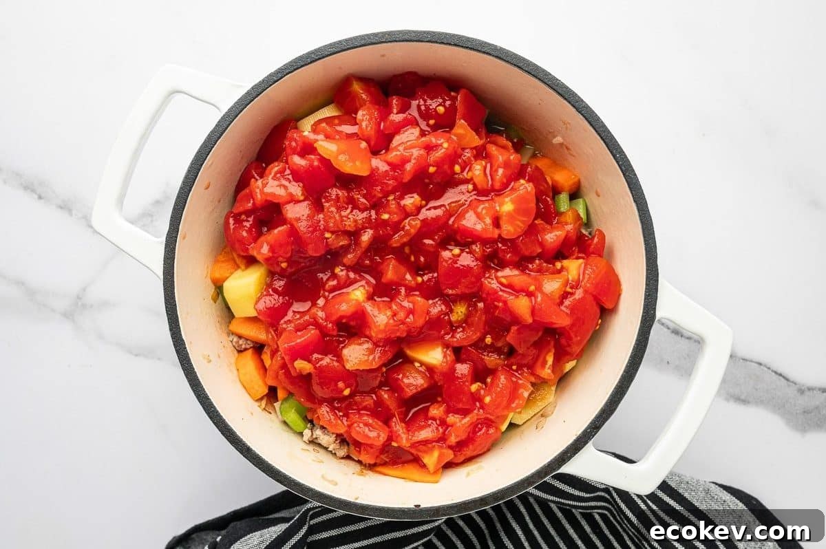 Overhead photo of canned diced tomatoes piled over carrots, potatoes, celery, and browned ground turkey in a white enameled Dutch oven.