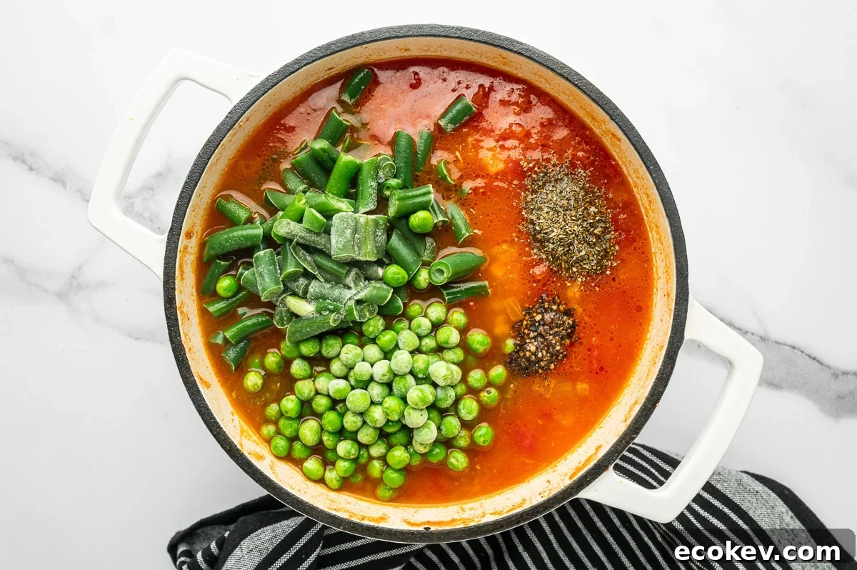 Overhead photo of red tomato broth in a white enameled Dutch oven topped with frozen green beans and peas, plus a mound of Italian seasoning and cracked black pepper.