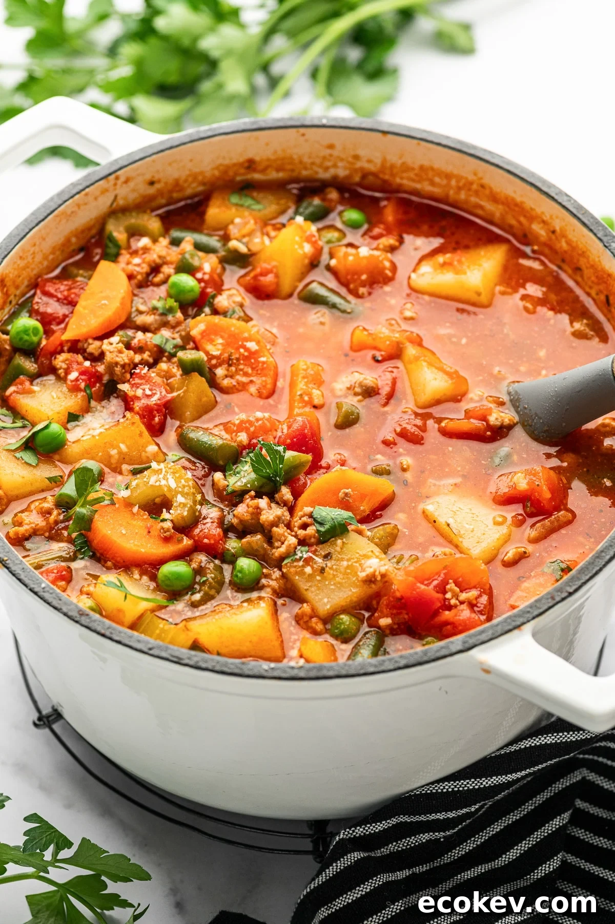 Close-up angled photo of ground turkey soup in a white enameled Dutch oven with a gray ladle resting in the red broth showing potatoes, carrots, green beans, peas, and crumbled turkey.