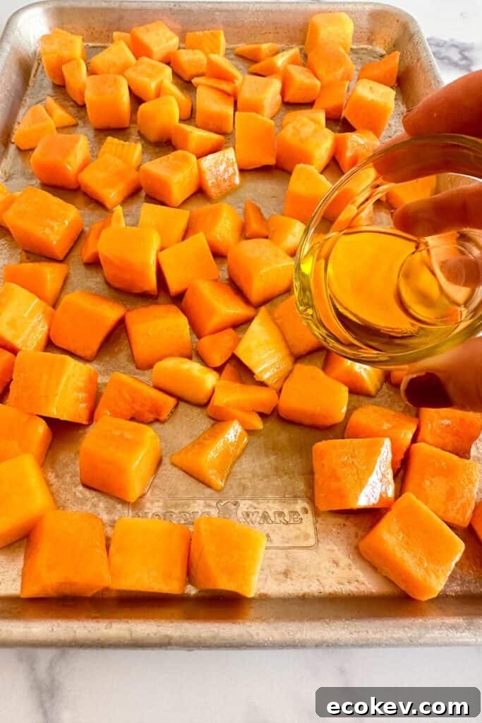 Olive oil being drizzled onto butternut squash cubes on a sheet pan.