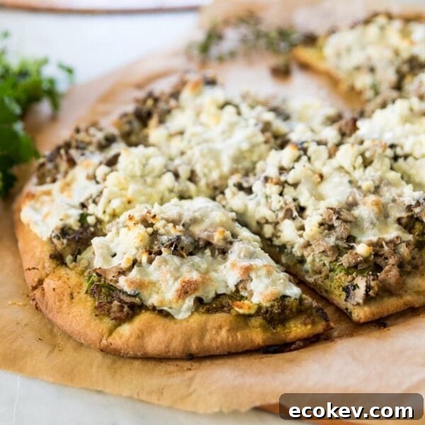 Homemade pizza crust topped with sausage, spinach, and crumbly feta cheese sitting on brown piece of parchment paper on marble surface after coming out of the oven