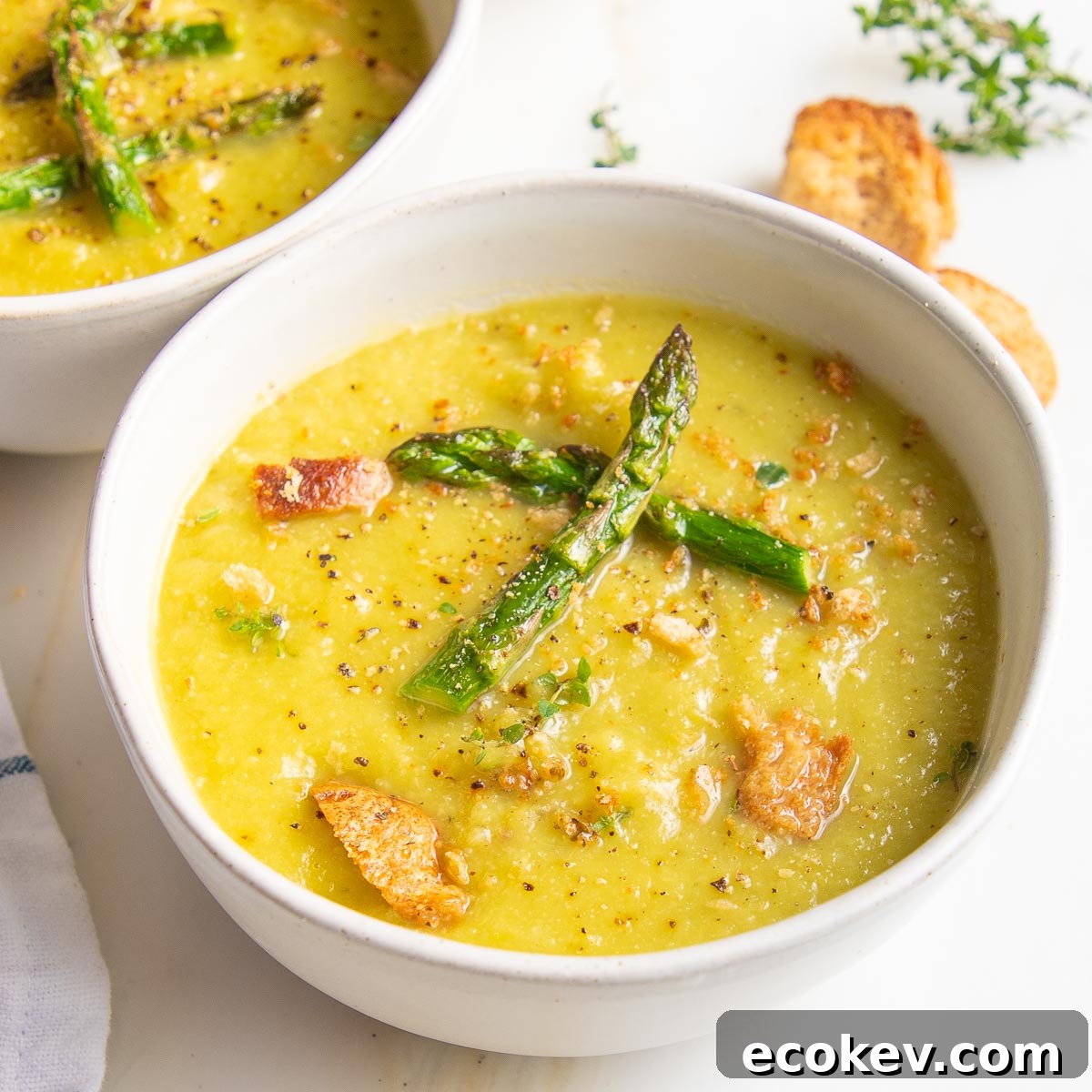 White bowl with yellowish-green thick soup, roasted asparagus tips floating in the center, and there is cracked black pepper and croutons sprinkled around.