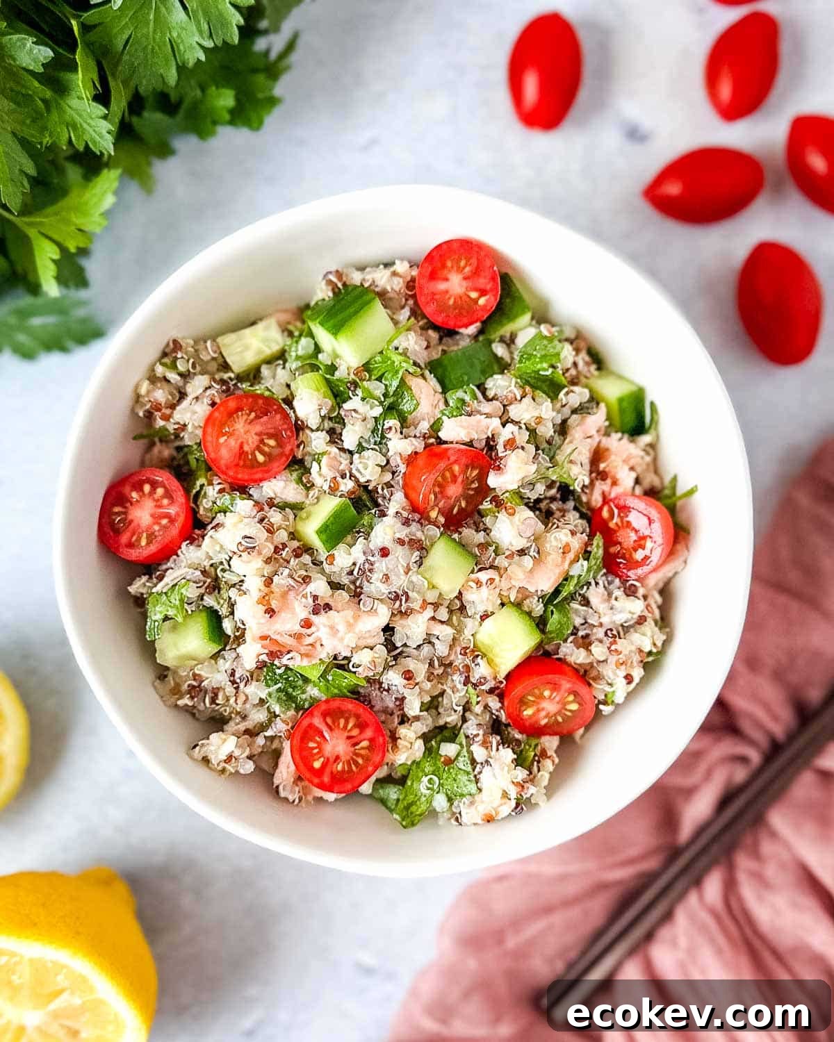Salmon quinoa salad in a white bowl surrounded by cherry tomatoes, fresh parsley, and a cut lemon.