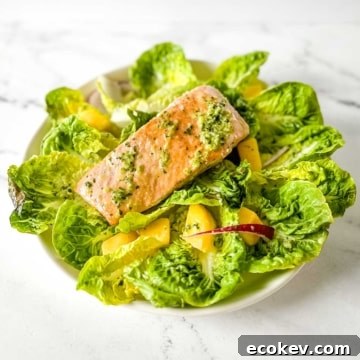 A fillet of salmon sits on a bed of little gem lettuce, mango, red onion, and jalapeno vinaigrette on a white marble counter.