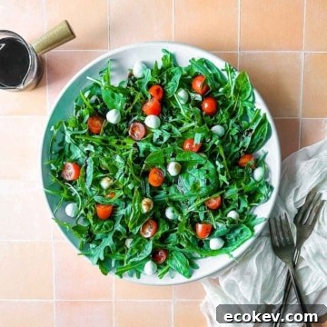 Arugula caprese salad on a white plate next to two forks, a white linen, and a small glass pitcher of balsamic glaze.