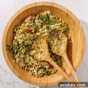 A wooden bowl filled with orzo pesto salad mixed with arugula, sun-dried tomatoes, feta cheese, and herbs, accompanied by two wooden serving utensils.
