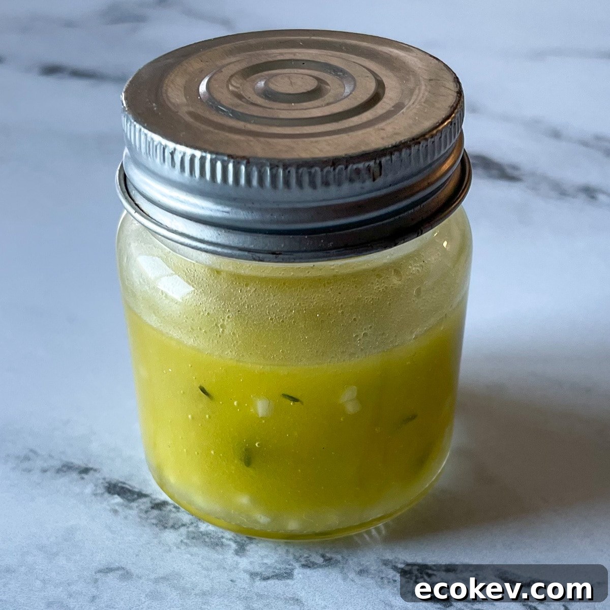 A small clear glass jar with a silver lid containing lemon, garlic, thyme vinaigrette sits on a marble counter top.