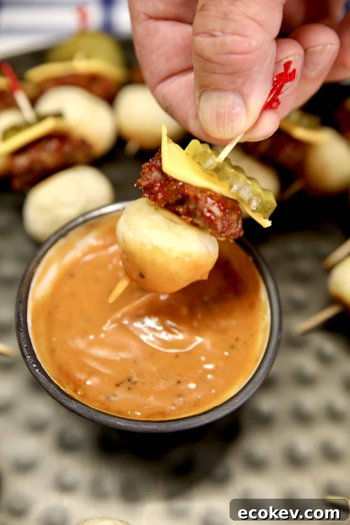 Mini cheeseburger appetizers on a stick, dipping into sauce.