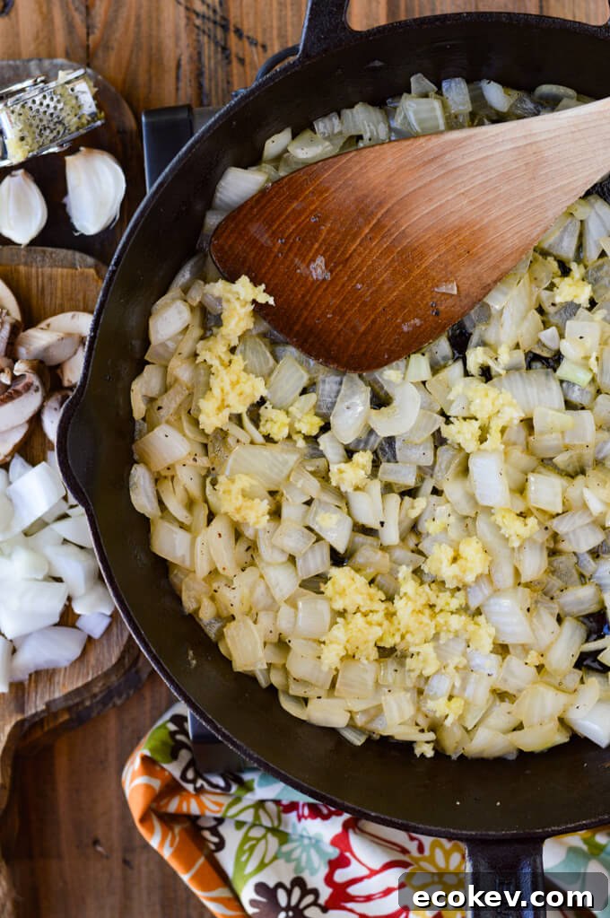 A cast iron skillet of sauteéd onions and minced garlic with raw mushrooms and chopped raw onions and a garlic press siitting on a wooden board.