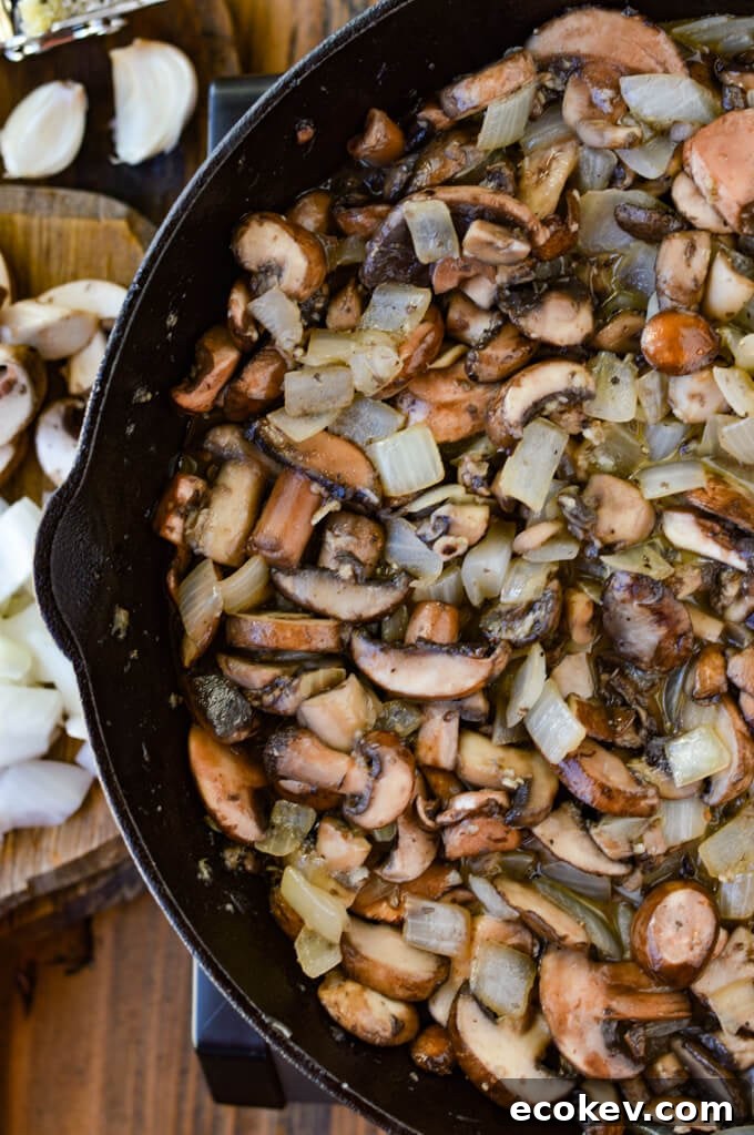 A cast iron skillet with sauteéd mushrooms and onions. A wooden board with raw chopped onions and raw mushrooms next to the pan.