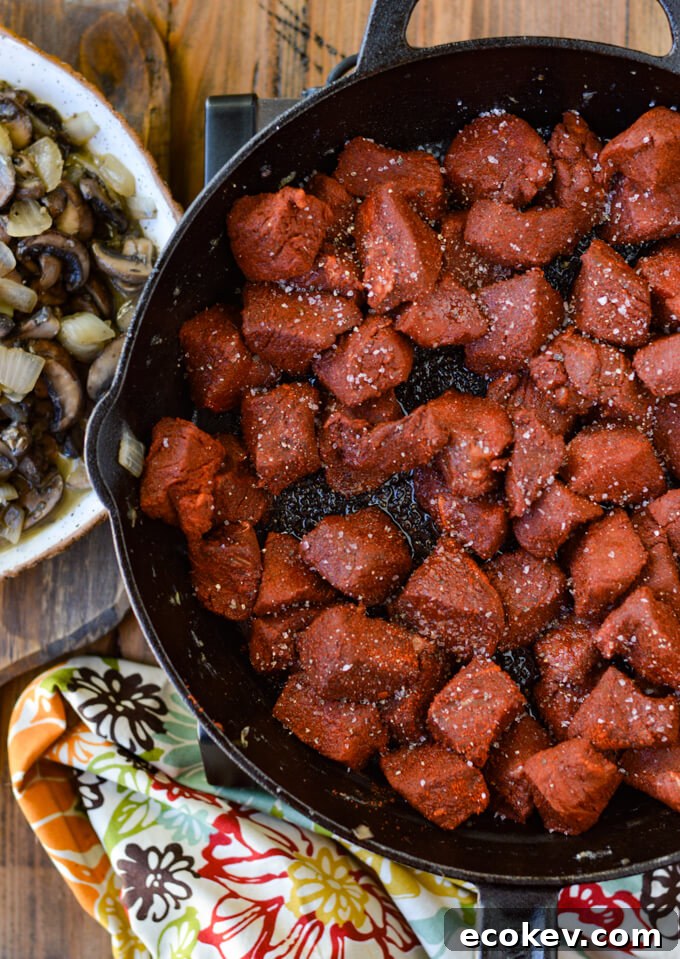 A cast iron skillet with chunks of raw sirloin steak coated with smoked paprika next to a dish of sauteéd mushrooms and onions and a colorful napkin.