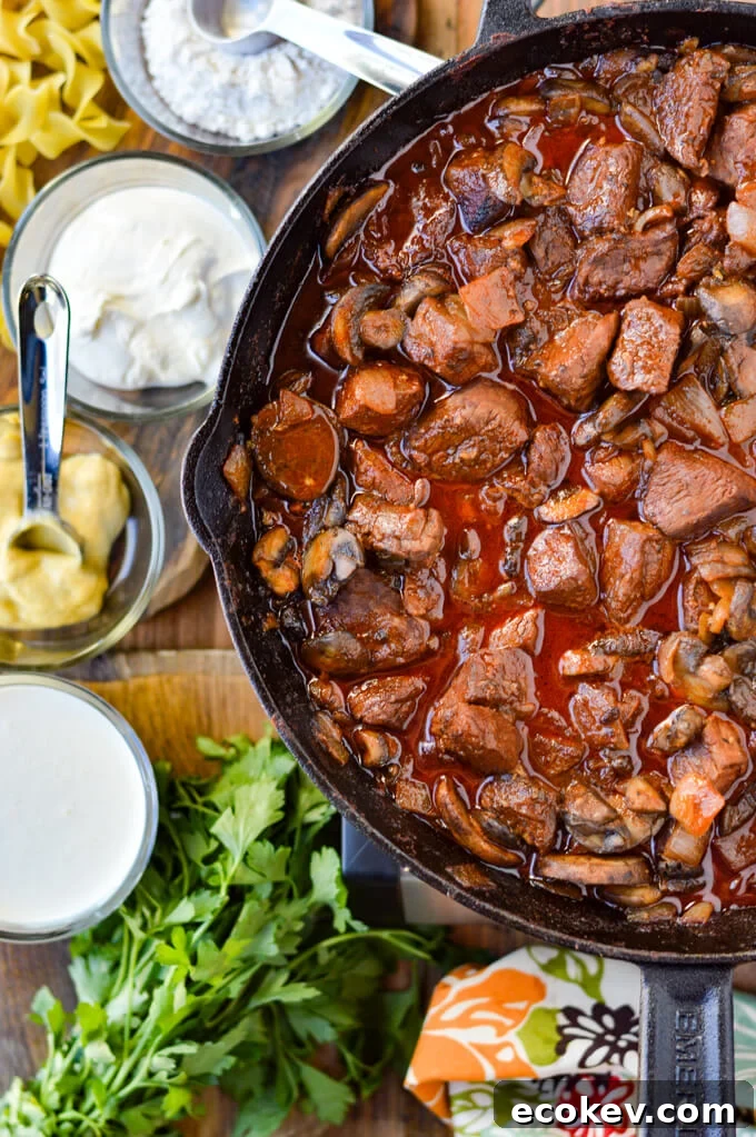 A cast iron skillet with beef stroganoff chunks. Sour cream, mustard, whole cream and stems of parsley are on a wooden board beside it.