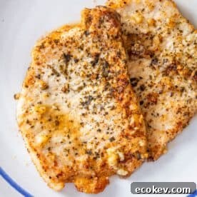 Two seasoned, cooked pork chops on a white plate, reminiscent of pan fried pork cutlet with garlic, sprinkled with herbs and spices.