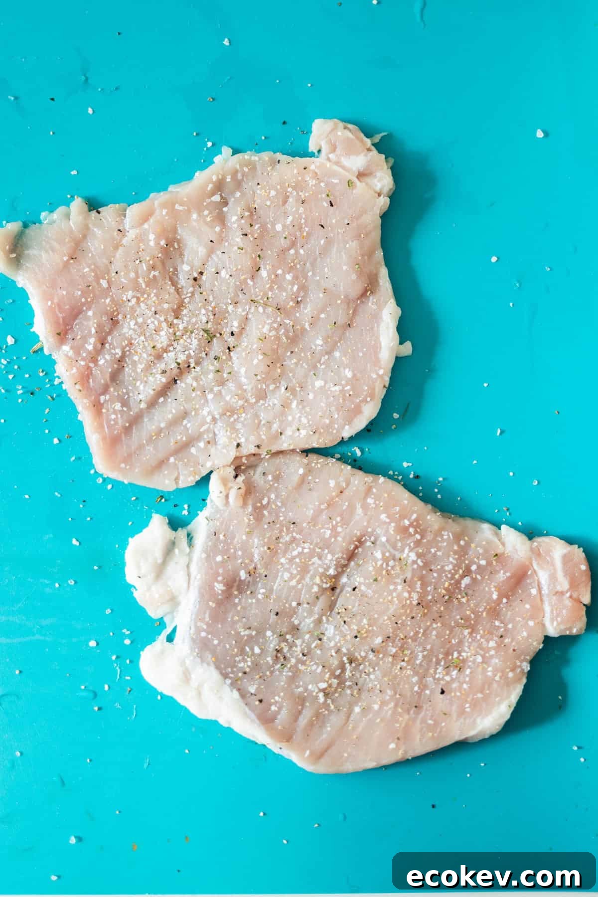 Two raw pork chops, perfect for a Pan Fried Pork Cutlet with Garlic, are seasoned with salt and pepper and placed on a bright turquoise cutting board.
