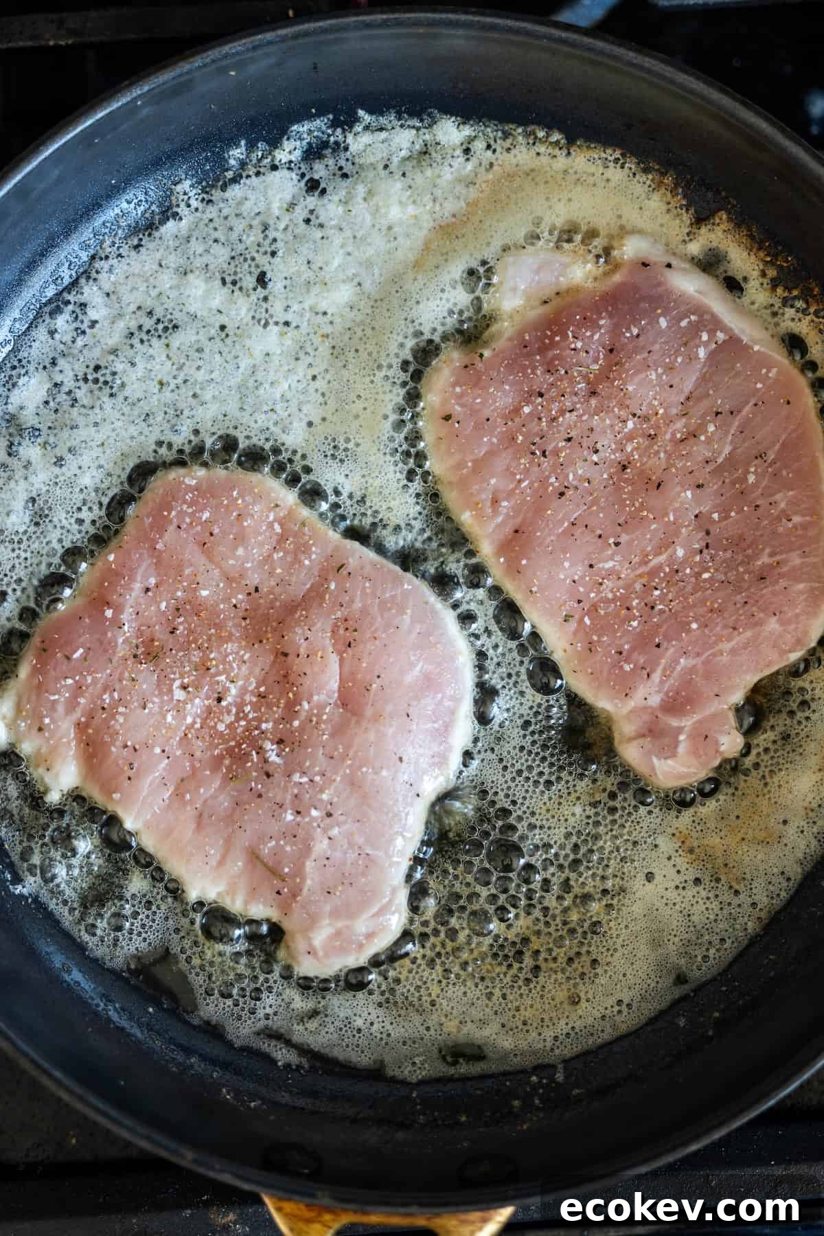 Two raw pork chops, perfect for a pan fried pork cutlet with garlic, are seasoned with salt and pepper as they sizzle in oil and butter in a frying pan.