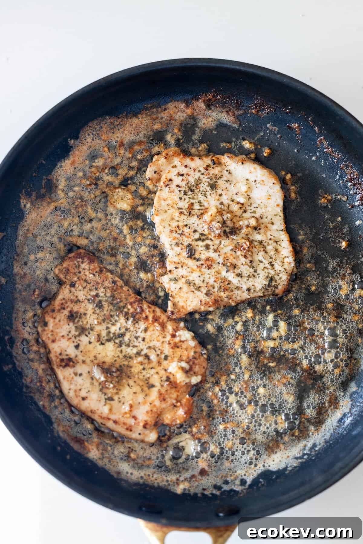 Two seasoned chicken breasts are being cooked in a black skillet, much like a Pan Fried Pork Cutlet with Garlic, with delicious browned bits and savory juices.