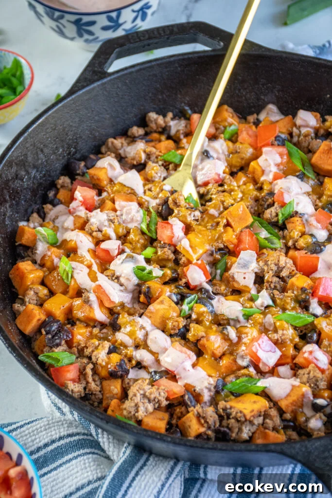 A gold serving spoon in a turkey and sweet potato skillet with black beans and garnished with diced tomatoes and a drizzle of sauce.