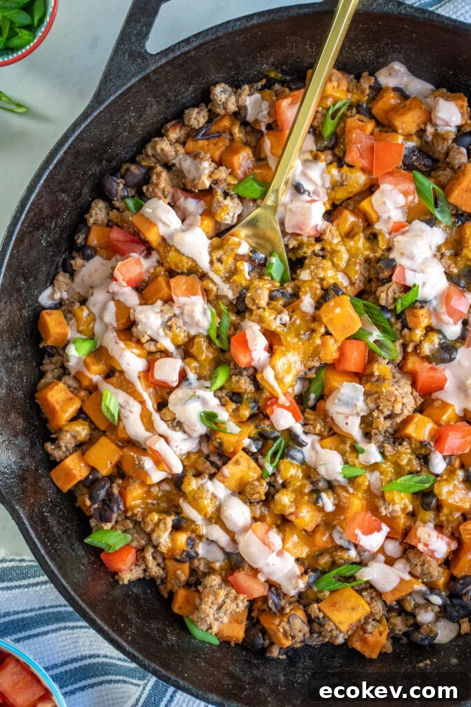 Close up of a cast iron skillet with ground turkey, sweet potatoes, and black beans, topped with diced tomatoes, green onions, and a sauce drizzle.