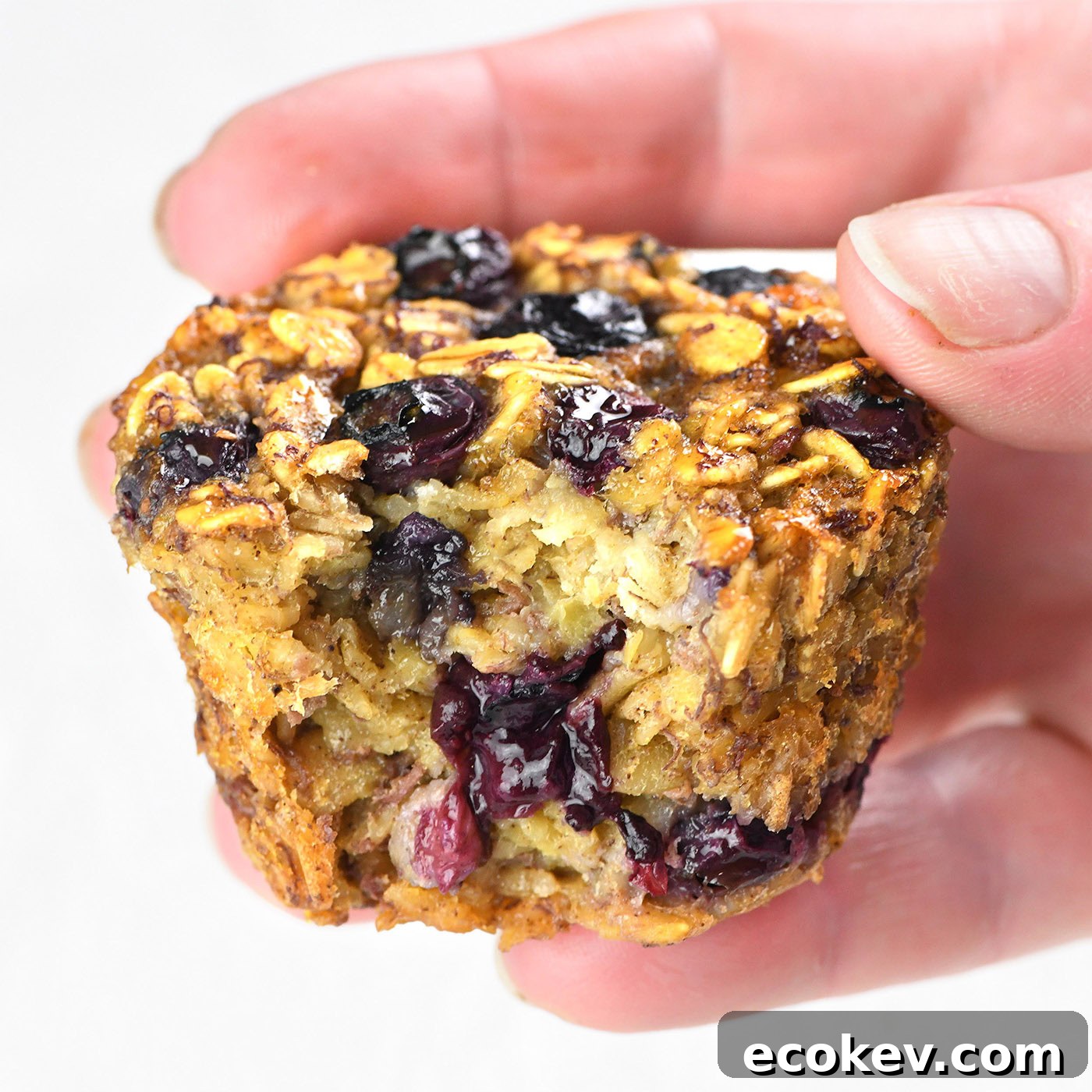 Hand holding a Blueberry Banana Baked Oatmeal Cup with a bite taken, showing the moist and fruity interior.