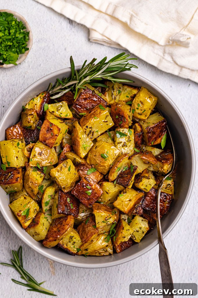 A bowl of roasted potatoes with a spoon sticking in the bowl. A sprig of rosemary is sticking out the side of the bowl.