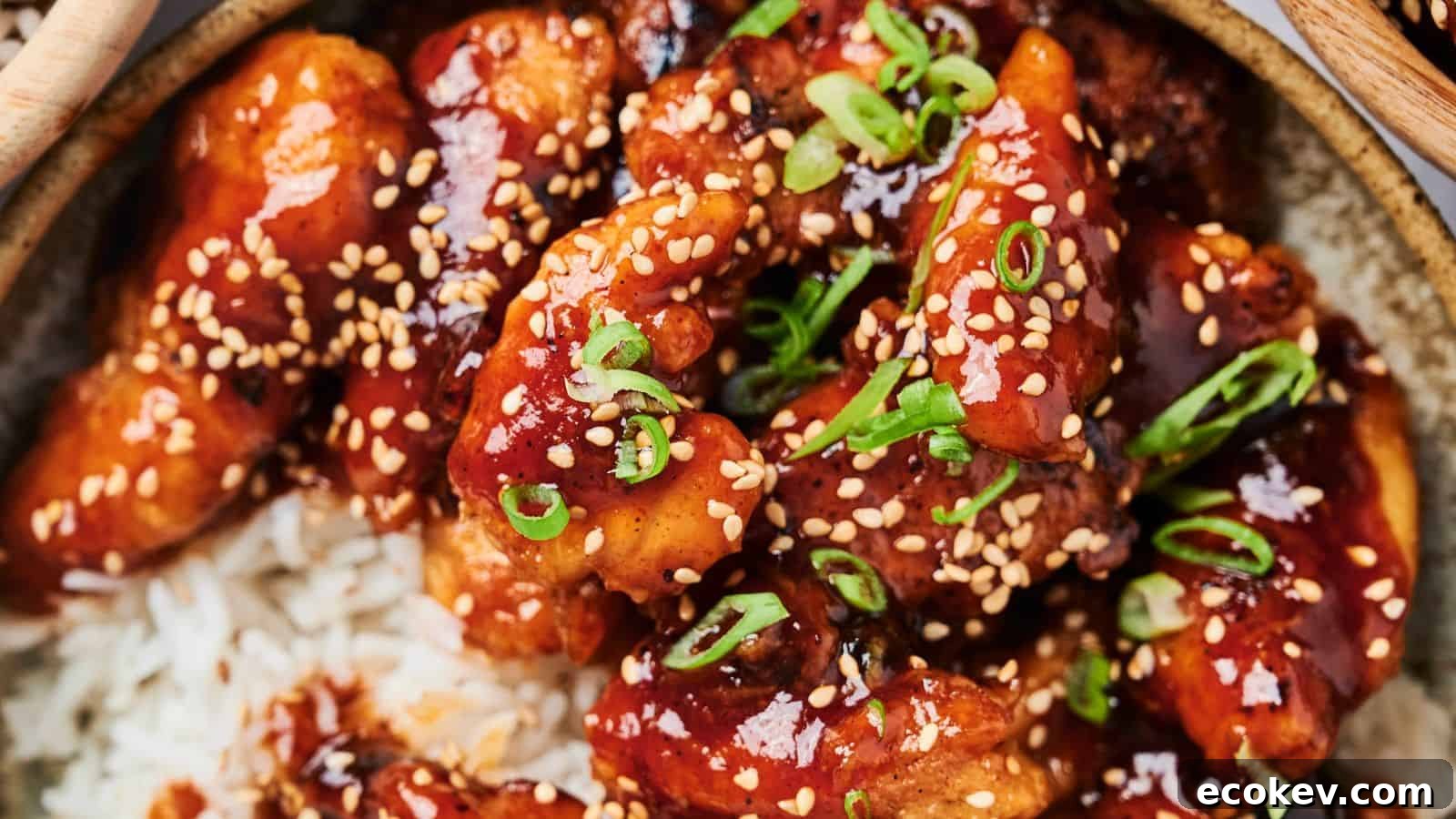A close-up of glazed chicken pieces with sesame seeds and chopped scallions on a bed of rice.