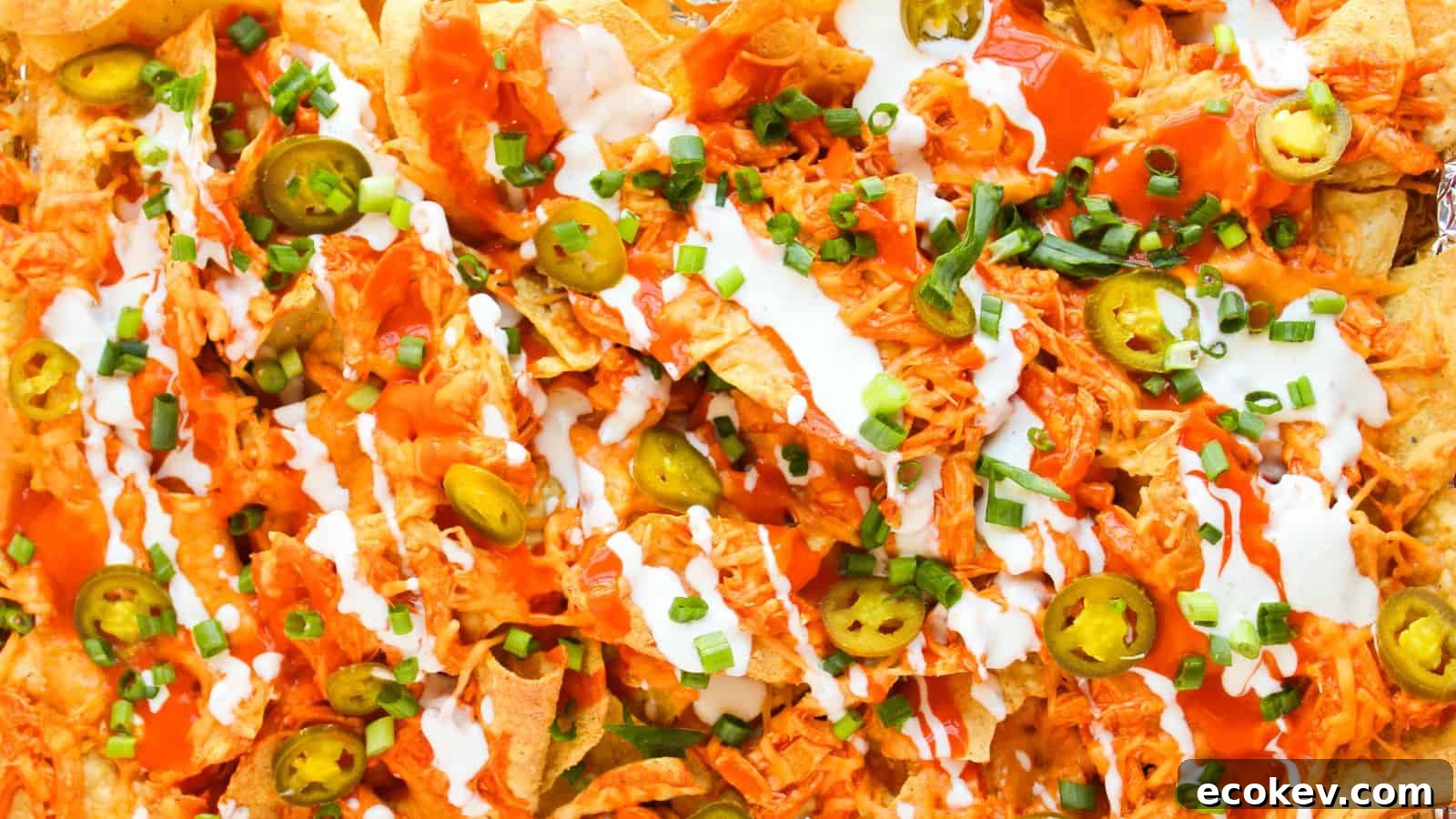 Close-up of loaded Buffalo chicken nachos piled high with shredded chicken, jalapeños, cheese, green onions, hot sauce, and a dollop of sour cream.