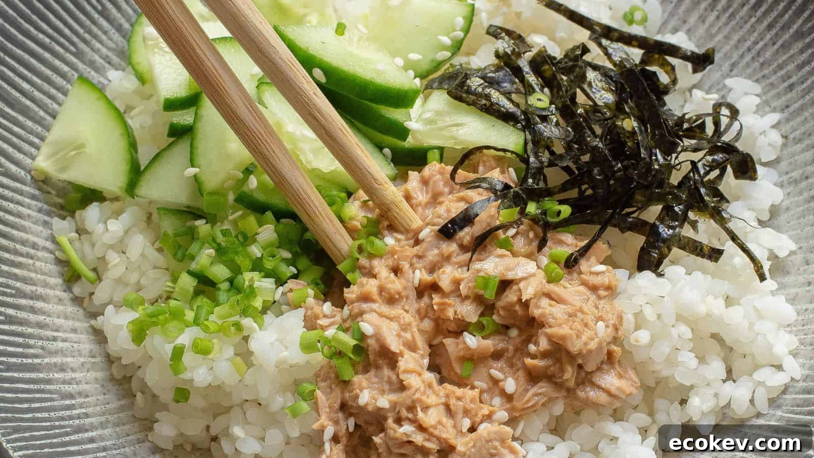 A bowl of white rice topped with sliced cucumber, shredded nori, chopped green onions, and a portion of tuna, with a pair of chopsticks resting on top.