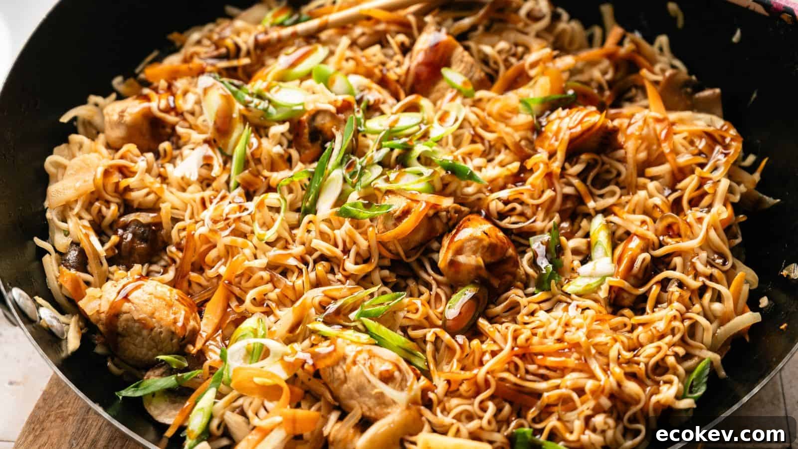 A close-up of cooked stir-fried noodles with pieces of chicken, sliced carrots, green onions, and a savory sauce in a black pan.