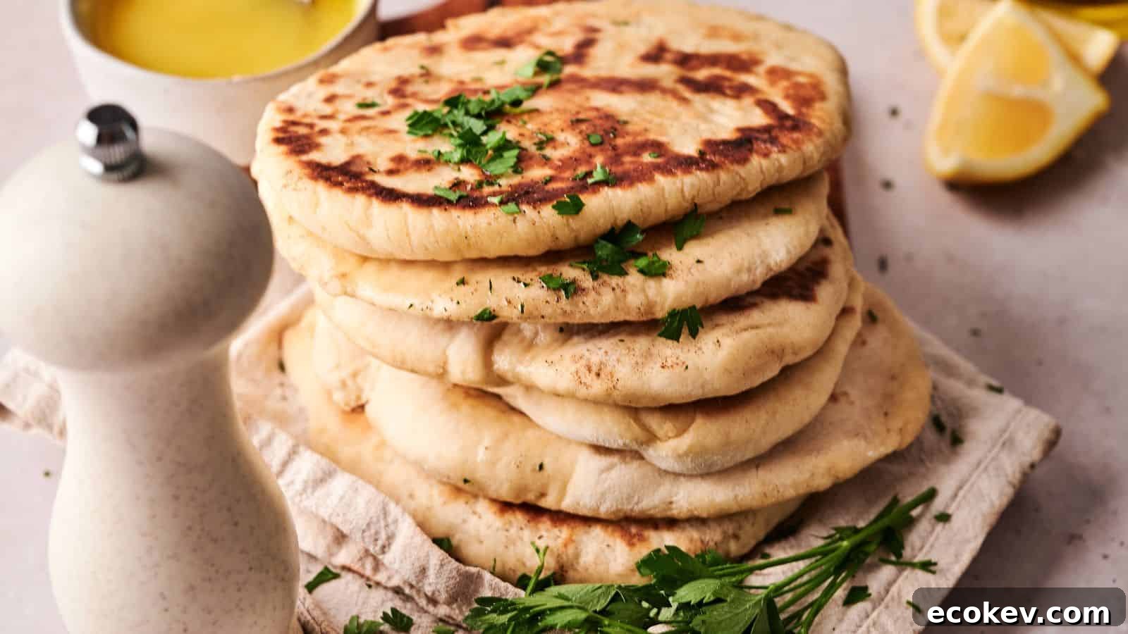 A stack of flatbreads garnished with chopped parsley sits on a cloth, with a white pepper grinder, fresh herbs, a lemon wedge, and a bowl of yellow sauce nearby.