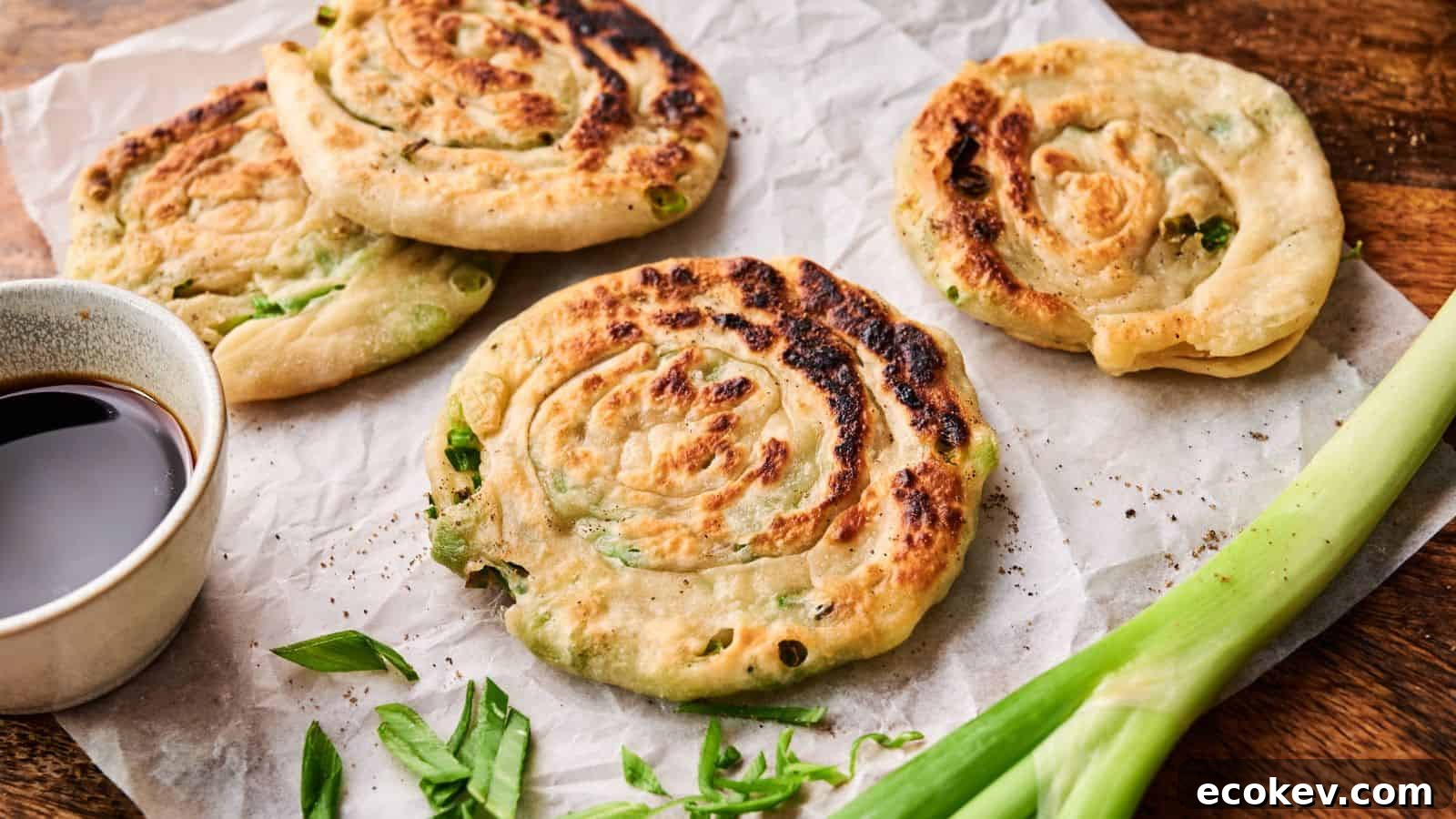 Four cooked scallion pancakes on parchment paper, garnished with sliced green onions, with a small bowl of dipping sauce and a whole scallion beside them.