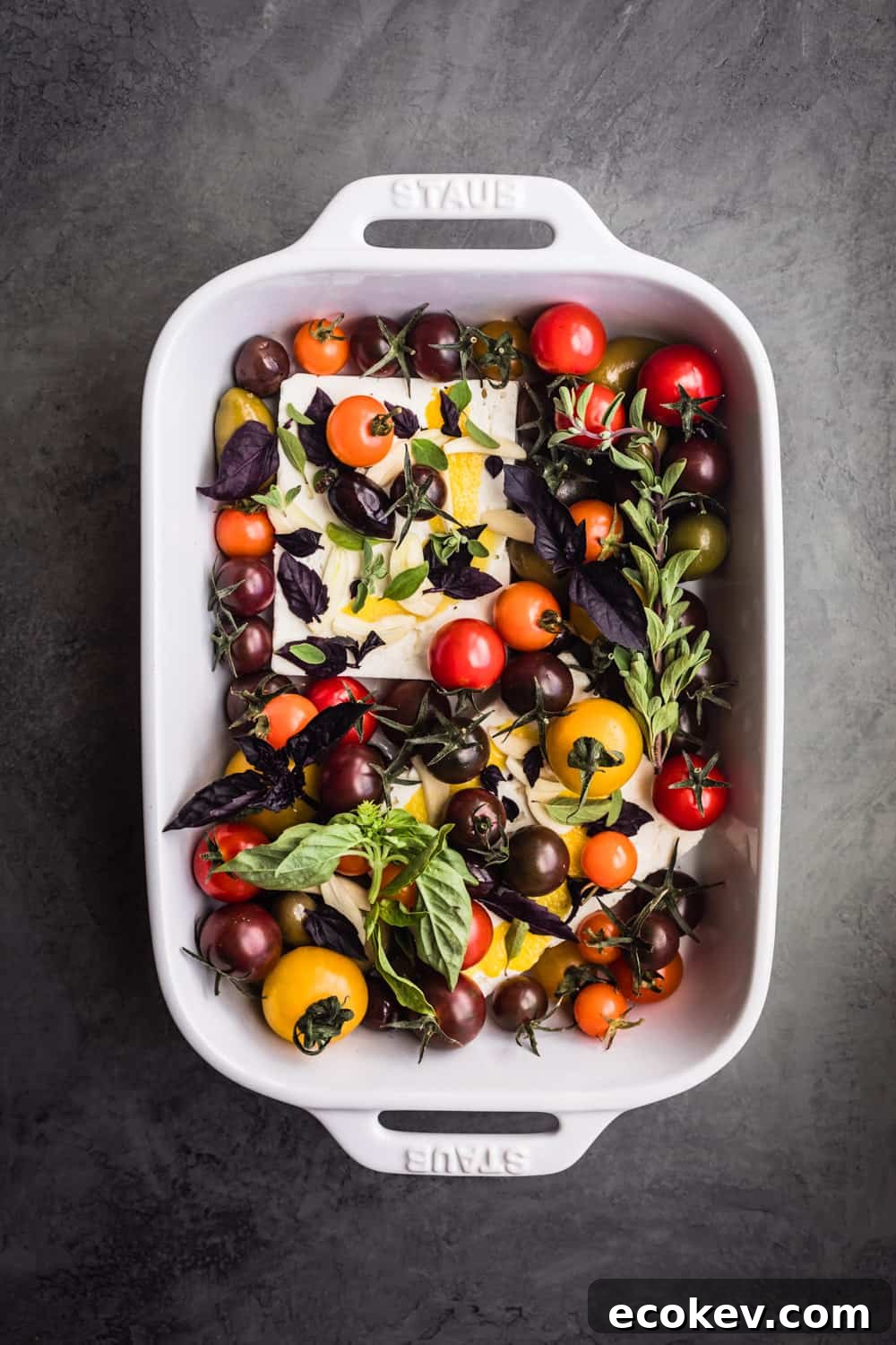 Feta in a baking dish with tomatoes, fresh herbs, lemon zest and garlic; overhead shot.