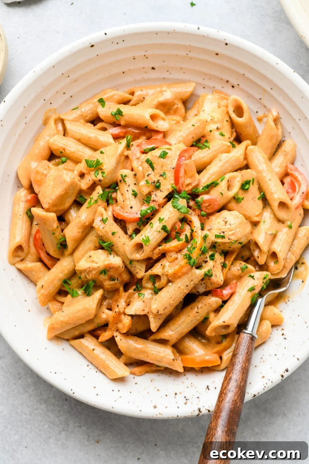 Large speckled shallow bowl filled with creamy cajun chicken penne pasta. Topped with a sprinkle of parsley and a wooden handled fork tucked into the side of the bowl. 