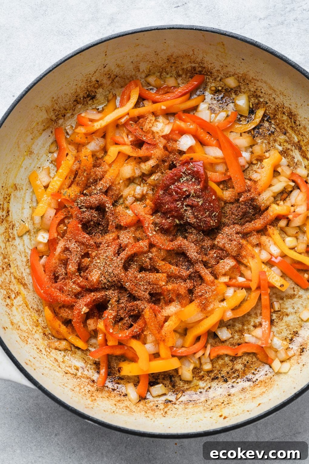 Cooked bell peppers, onion, and garlic, in a large white ceramic skillet with tomato paste and spices on top.
