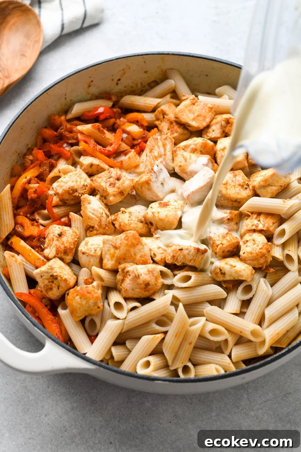 Cooked veggies, chicken, and pasta in a large white ceramic skillet with cashew cream sauce being poured in from a blender container.