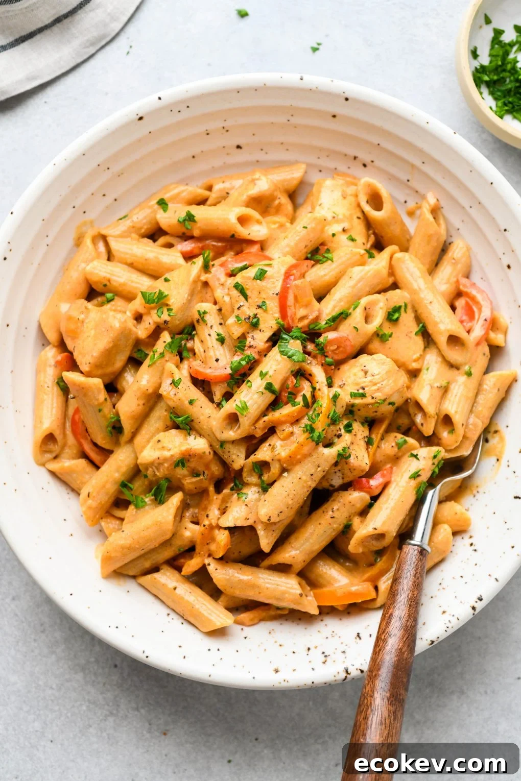 Large speckled shallow bowl filled with creamy cajun chicken penne pasta. Topped with a sprinkle of parsley and a wooden handled fork tucked into the side of the bowl. 