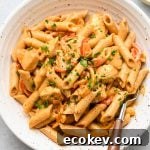 A bowl of creamy penne pasta with sliced red bell peppers and sprinkled chopped parsley, served with a fork on the side.