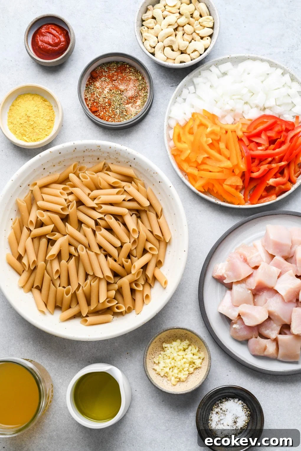 Ingredients for cajun chicken pasta on a light colored background. A bowl of cajun spice blend, a bowl of nutritional yeast, a bowl of chopped garlic, a bowl of uncooked penne pasta, a plate with chopped onion and bell peppers, and a plate with diced raw chicken.