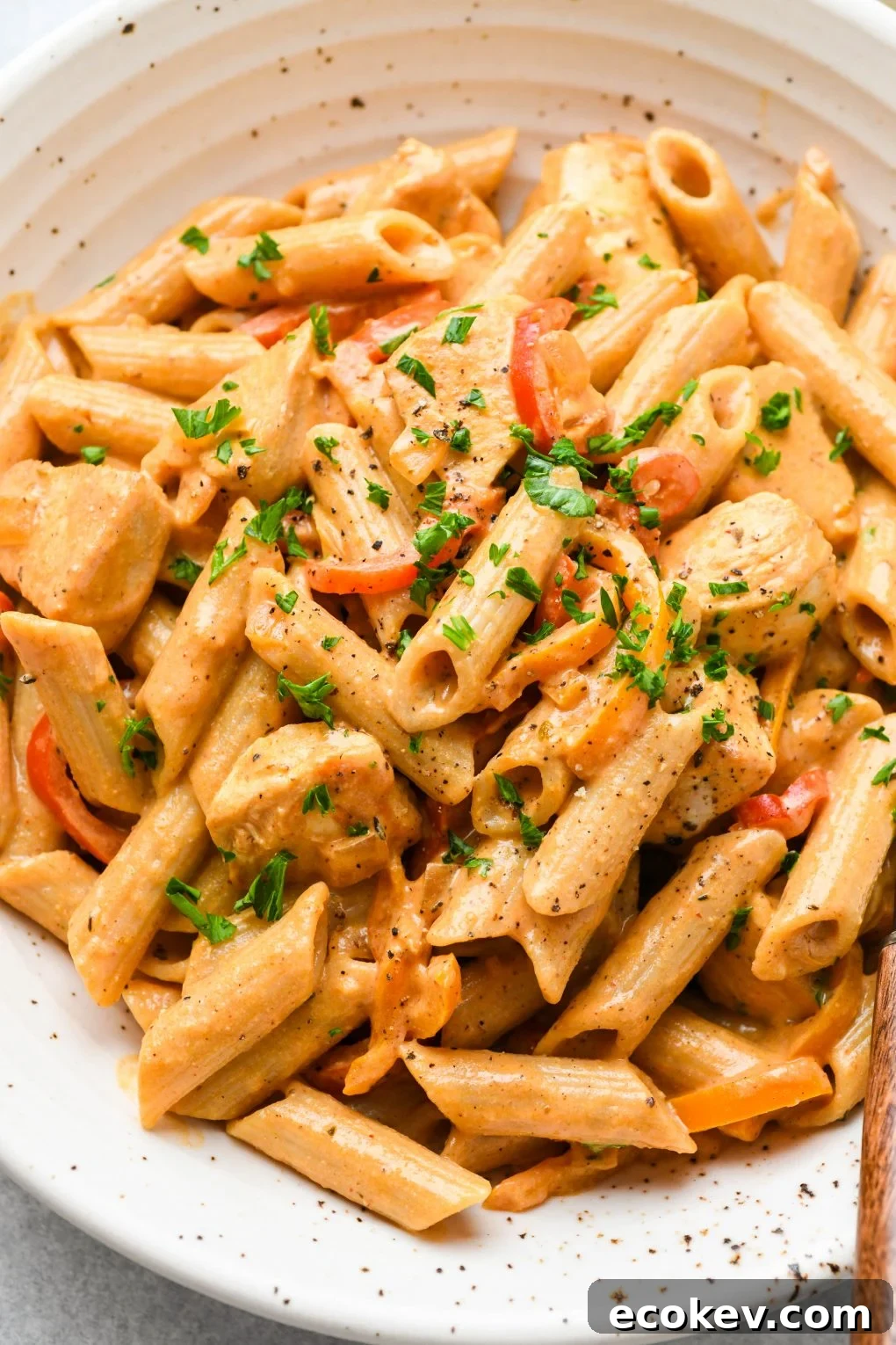 Close up shot of a large speckled shallow bowl filled with creamy cajun chicken penne pasta. Topped with a sprinkle of parsley and a wooden handled fork tucked into the side of the bowl. 