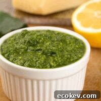A small white bowl filled with kale pesto with a half lemon and cutting board with a wedge of Parmesan cheese in the background.