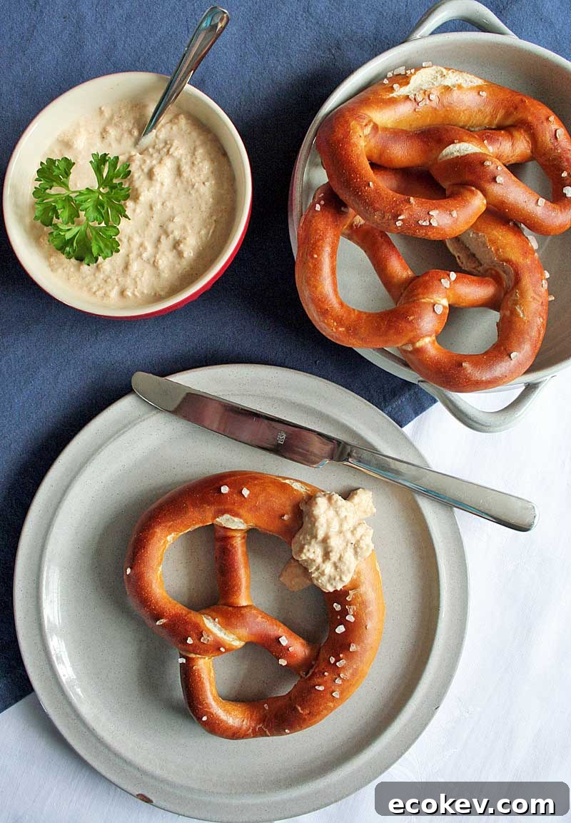 Top down view of three large lye pretzels on platters with a bowl of German Obatzda Bavarian Cheese Dip on a blue table cloth.