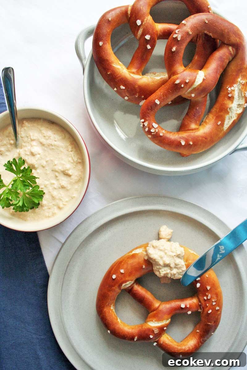 Top down view of a three lye pretzels on white, porcelain plates with a a bowl of German Obatzda cheese dip placed to the left.