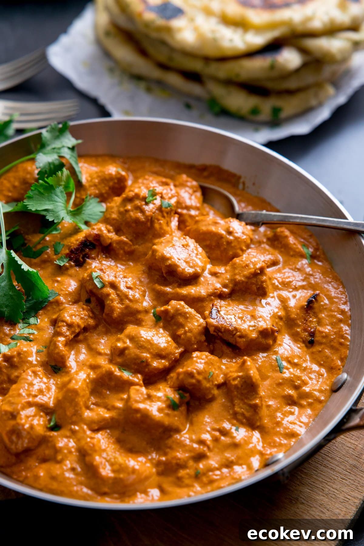 Silver frying pan with butter chicken curry in it. Pile of naan bread in the background.