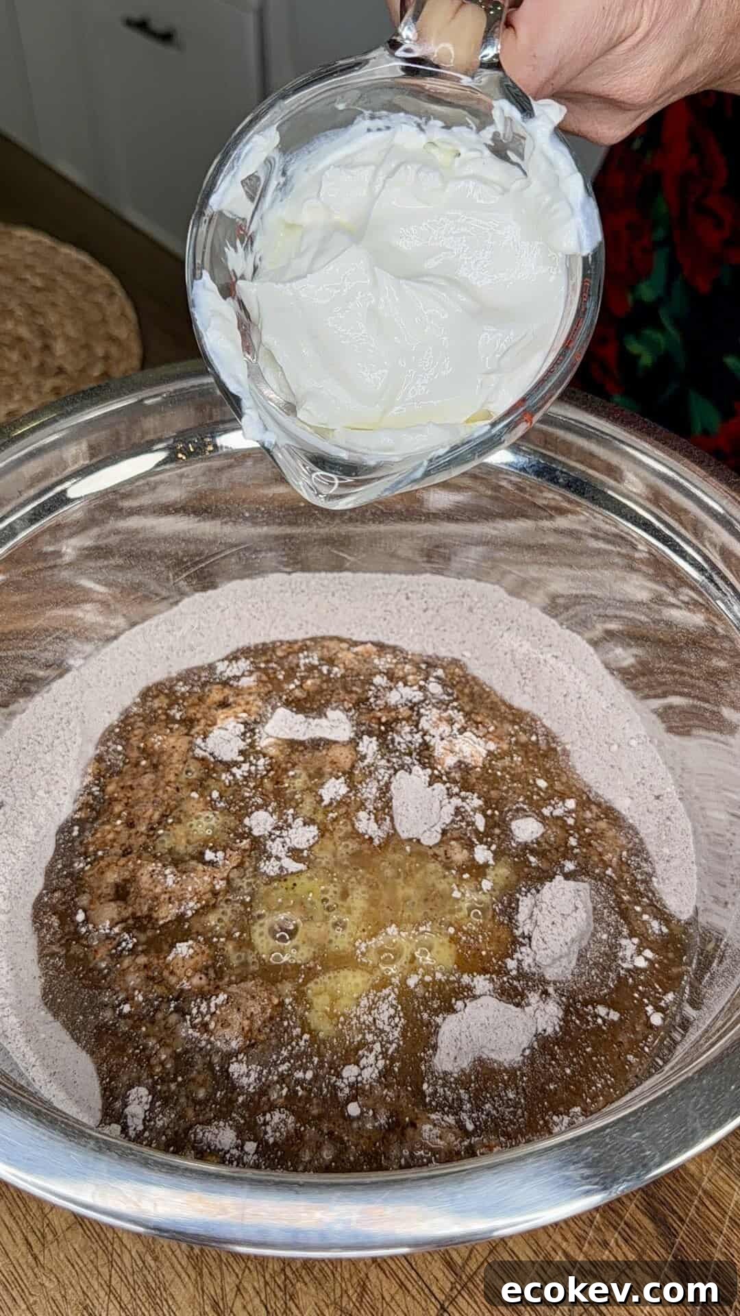 A hand holds a bowl of thick white yogurt above a metal mixing bowl containing a blend of dry and wet baking ingredients on a wooden countertop.